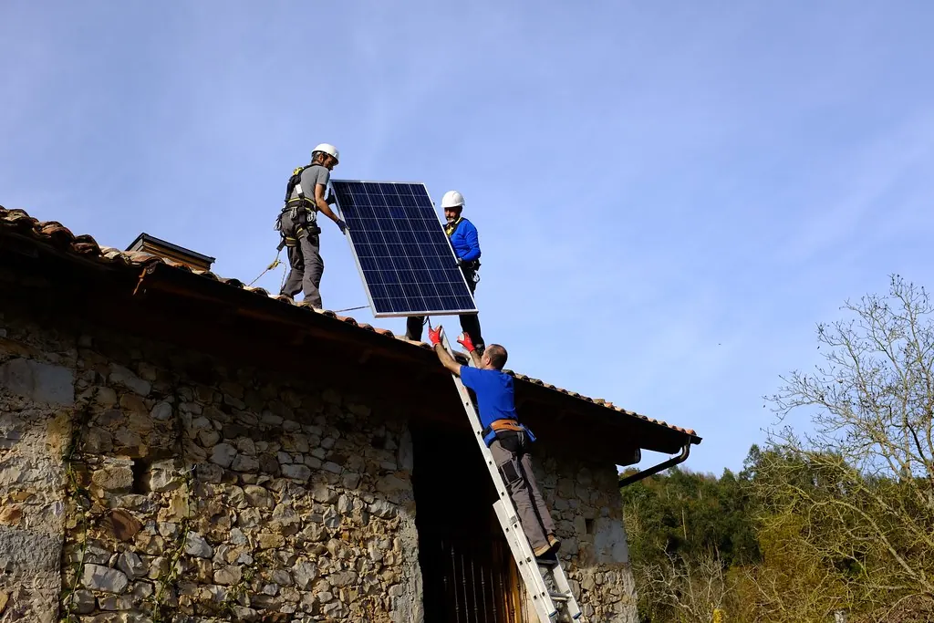Three installers lifting a solar panel onto the tiled roof of a stone house